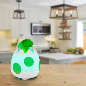 May include: An egg-shaped essential oil diffuser with a green dinosaur emerging from the top. The diffuser is white with green polka dots and a light blue glow. The dinosaur is bright green, and vapor is emitted from its mouth.