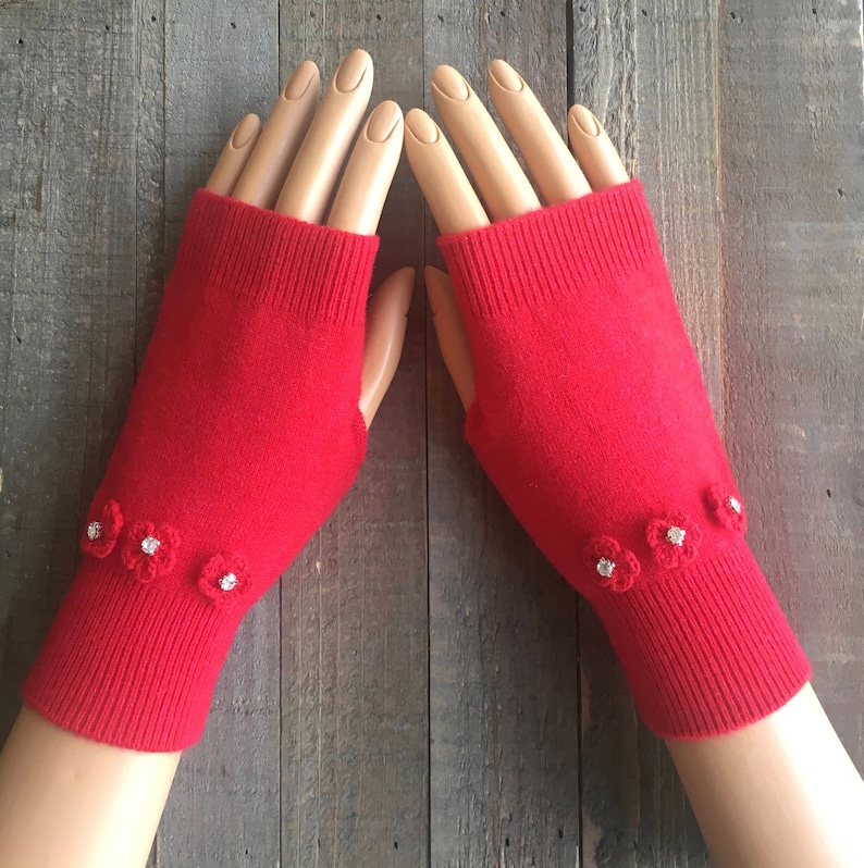 May include: Red fingerless gloves with white flower embellishments. The gloves are made of a soft, knitted fabric.