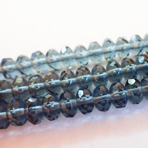 May include: Close-up of three strands of faceted, blue-gray beads. The beads are strung together, with each bead displaying multiple facets that catch the light. The background is a plain white.