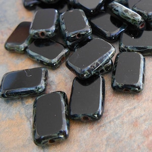 May include: A collection of rectangular black glass beads with rounded corners. The beads have a glossy finish and small holes for stringing. Some beads show a subtle iridescent green and blue pattern.