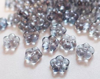 May include: A collection of translucent, flower-shaped beads in shades of gray and blue. The beads are clustered together, with several in sharp focus, showcasing their delicate, five-petal design. These beads are ideal for jewelry making and crafting projects.