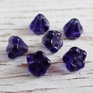 May include: Six dark purple, flower-shaped glass beads are scattered on a white, distressed wooden surface. The beads have a glossy finish and a unique, textured design. The beads are approximately 0.75 inches in diameter, perfect for jewelry making or craft projects.