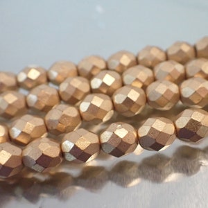 May include: Close-up of strands of faceted, metallic rose gold beads. The beads have a geometric, multi-faceted design, reflecting light. The beads are strung together, suggesting their use in jewelry making or crafting projects.
