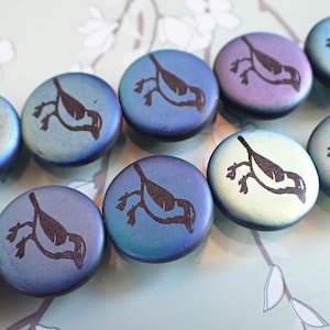 May include: Close-up of several round beads with a metallic finish in shades of blue, purple, and green. Each bead features a dark brown bird design. The beads are arranged closely together, showcasing their iridescent quality.