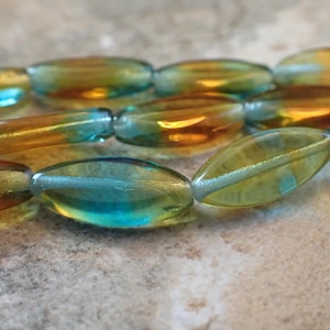 May include: Close-up of a strand of oval-shaped beads. The beads have a translucent quality with a blend of colors, including teal, gold, and amber. The beads are strung together, suggesting they are for jewelry making or crafting.