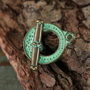 May include: A close-up of a toggle clasp with a bronze finish and a turquoise patina. The clasp features a circular ring with a textured surface and a bar that slides through it. The clasp is set against a textured, brown background.