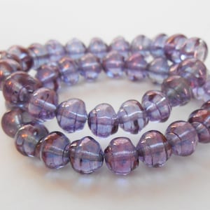 May include: Close-up of a strand of purple glass beads. The beads are round with a ribbed texture and a translucent quality. The color shifts from a deep purple to a lighter lavender, with hints of iridescence. The beads are strung together, forming a circular shape.