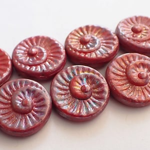 May include: A collection of red, iridescent, spiral-shaped beads. Each bead features a central raised point and a detailed, textured surface. The beads are closely arranged against a white backdrop, showcasing their unique design and colour.