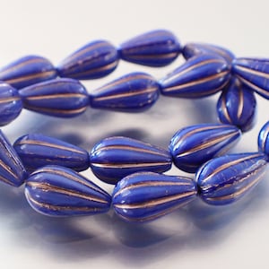 May include: A close-up shot of a string of blue, teardrop-shaped beads with gold stripes. The beads are arranged in a curved pattern, showing their glossy finish and detailed design. The background is a soft, neutral tone, highlighting the vibrant blue and gold.