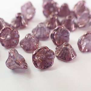 Puede incluir: Primer plano de varias cuentas translúcidas moradas con forma de flor. Las cuentas tienen una superficie texturizada y un agujero en el centro, ideal para ensartar. Las cuentas están dispersas sobre una superficie blanca.