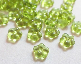 May include: A collection of translucent, light green flower-shaped beads. The beads have five petals and a small hole in the center. The beads are scattered across a white surface, with some in focus and others blurred.