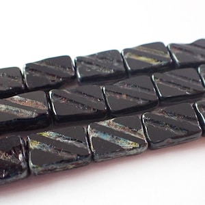 May include: Close-up of a strand of black rectangular beads with diagonal etched lines. The beads have a glossy finish and iridescent accents, suggesting they are made of glass or a similar material. The beads are arranged in a row.
