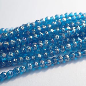 May include: Close-up of strands of faceted, light blue beads. The beads have a metallic, silver-like finish on their facets, creating a sparkling effect. The beads are strung together, suggesting their use in jewelry making or crafts.