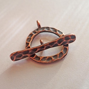 May include: A copper-colored toggle clasp, featuring a textured, hammered design. The clasp consists of a circular ring with a small loop and a bar that fits through the ring. Ideal for jewelry making.