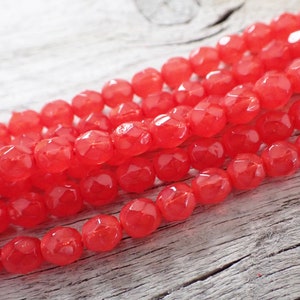 May include: Strands of faceted, bright red beads. The beads have a glossy finish and are arranged diagonally across a light-colored, textured wooden surface. The beads are likely for jewelry making.