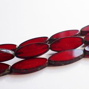 May include: Close-up of a collection of oval-shaped, deep red glass beads. The beads have a glossy finish and a dark, almost black, border. The beads are arranged in a diagonal pattern against a white background.