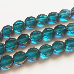 May include: Close-up of three strands of faceted teal beads. The beads have a metallic bronze luster on the edges, and a deep teal color in the center. The beads are round with multiple facets.