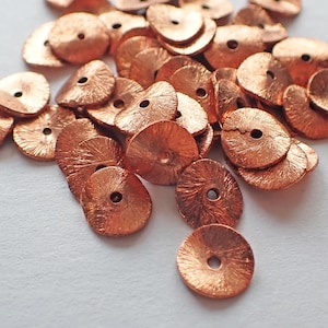 May include: A collection of copper-coloured, textured metal sequins. Each sequin is round with a central hole and a slightly domed shape. The sequins are scattered, creating a visually appealing texture.