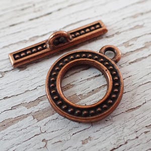 May include: Close-up of two antique copper-colored jewelry clasps. One is a circular toggle clasp with a dotted border, and the other is a rectangular bar with a loop. The clasps are on a distressed white wooden surface.
