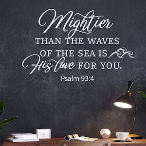 Puede incluir: Una pared oscura con una inscripción blanca que dice: "Más poderoso que las olas del mar es su amor por ti. Salmo 93:4." Un escritorio de madera con una lámpara, libros y una taza de té está frente a la pared. Hay plantas verdes a ambos lados.