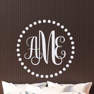 Może przedstawiać: Brązowe łóżko z białymi poduszkami i białą naklejką z monogramem na ścianie nad zagłówkiem. Monogram znajduje się w kółku z białymi kropkami wokół krawędzi. Litery to "AME".