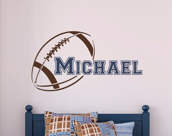 Könnte beinhalten: Ein brauner Football mit weißen Schnallen und einem blauen Namen "MICHAEL" an einer weißen Wand. Der Football ist ein Wandaufkleber.