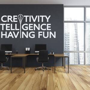 May include: A modern office space with two black desks and chairs. The wall is painted gray and features a white vinyl decal with the text "Creativity is Intelligence Having Fun" and a lightbulb icon.