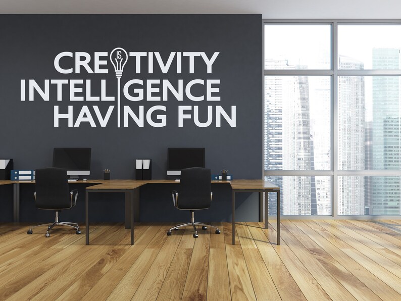 May include: A modern office space with two black desks and chairs. The wall is painted gray and features a white vinyl decal with the text "Creativity is Intelligence Having Fun" and a lightbulb icon.