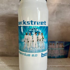 Puede incluir: Un vaso cilíndrico blanco con tapa y base metálicas, que presenta un gráfico de cinco personas con trajes blancos sobre un fondo digital azul. El texto "backstreet" aparece sobre la imagen, con "millennium 2.0 boy" impreso debajo.