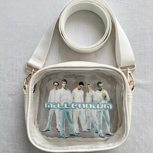 Puede incluir: Bolso bandolera color crema con un panel frontal transparente que muestra un grupo de personas con trajes blancos. La palabra "Millennium" está impresa en azul y plata. El bolso tiene una correa blanca y herrajes dorados.