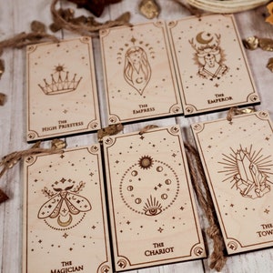 May include: Set of ten wooden tarot cards with gold foil accents and string for hanging. The cards feature illustrations of the High Priestess, Empress, Emperor, Magician, Chariot, and Tower.