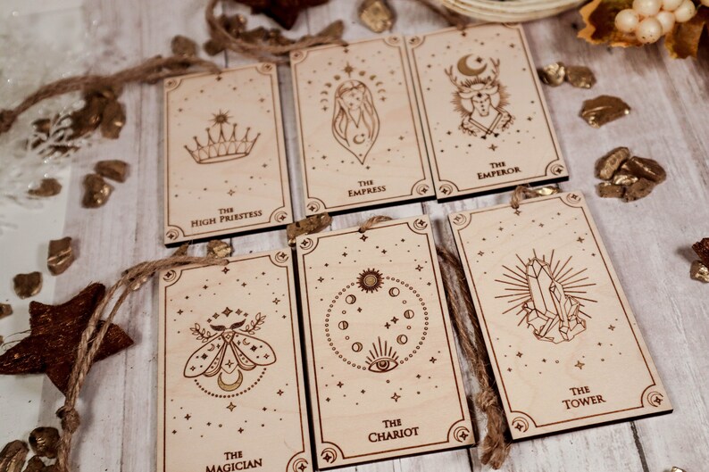 May include: Set of ten wooden tarot cards with gold foil accents and string for hanging. The cards feature illustrations of the High Priestess, Empress, Emperor, Magician, Chariot, and Tower.