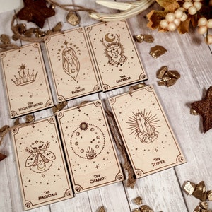 May include: Seven wooden ornaments featuring tarot card imagery. The cards are The High Priestess, The Empress, The Emperor, The Chariot, The Tower, The Magician, and The Star. Each card has a unique design and is decorated with gold glitter.