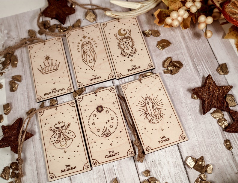 May include: Seven wooden ornaments featuring tarot card imagery. The cards are The High Priestess, The Empress, The Emperor, The Chariot, The Tower, The Magician, and The Star. Each card has a unique design and is decorated with gold glitter.