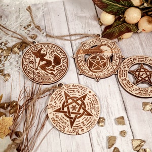 May include: Four wooden ornaments with a witch, a pentagram, and a phrase about surviving the darkness. The ornaments are hanging from a twine string and are on a white wooden background with gold glitter.