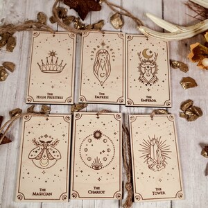 May include: Set of seven wooden tarot cards with gold foil accents. The cards feature illustrations of the High Priestess, Empress, Emperor, Magician, Chariot, Tower, and a moth. Each card has a star-studded background.