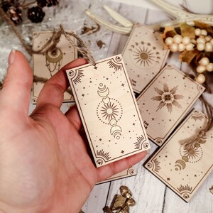 May include: A set of six wooden gift tags with celestial designs. Each tag features a different celestial motif, including a sun, moon, and stars. The tags are made of natural wood and have a rustic, bohemian aesthetic.