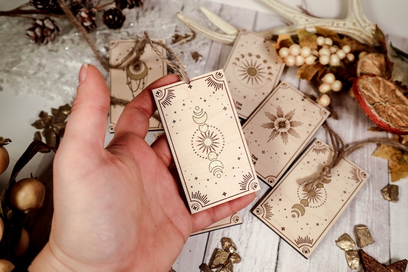 May include: A set of six wooden gift tags with celestial designs. Each tag features a different celestial motif, including a sun, moon, and stars. The tags are made of natural wood and have a rustic, bohemian aesthetic.