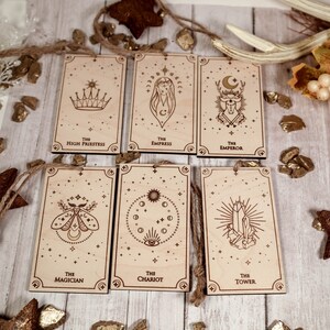 May include: Six wooden tarot cards with gold foil accents and twine hanging loops. The cards feature illustrations of the Magician, High Priestess, Empress, Emperor, Chariot, and Tower.