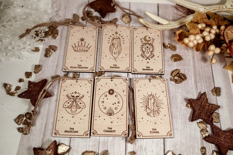 May include: Six wooden tarot cards with gold foil accents and twine hanging loops. The cards feature illustrations of the Magician, High Priestess, Empress, Emperor, Chariot, and Tower.