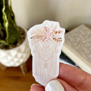 May include: A sticker featuring a bohemian-style design. The sticker has a floral arrangement in shades of pink and beige at the top, with a macrame-style pattern below. The sticker is held by a person.