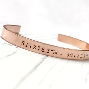 Puede incluir: Una pulsera de metal de color oro rosa con las coordenadas "51.2763° N, 30.2219° E" grabadas en ella.