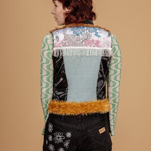 May include: A black, blue, and white vest with a fur trim and a floral pattern. The vest is worn over a green and white patterned long-sleeved shirt. The person is wearing black jeans with a white daisy design on the back pocket.