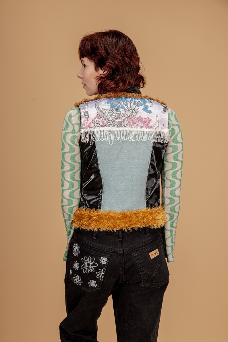 May include: A black, blue, and white vest with a fur trim and a floral pattern. The vest is worn over a green and white patterned long-sleeved shirt. The person is wearing black jeans with a white daisy design on the back pocket.
