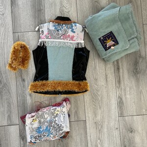May include: A blue and black vest with a white and pink patterned design, a brown fur trim, and a white fringe. The vest is hanging on a white hanger. There is a brown fur muff and a blue blanket with a sun design and the text "Solar Bell" in the image.