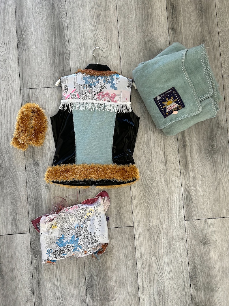 May include: A blue and black vest with a white and pink patterned design, a brown fur trim, and a white fringe. The vest is hanging on a white hanger. There is a brown fur muff and a blue blanket with a sun design and the text "Solar Bell" in the image.