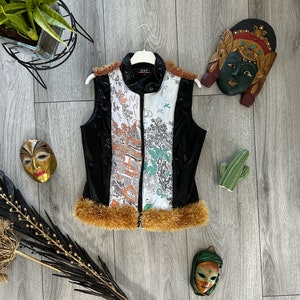 May include: Black sleeveless vest with a white and orange patterned design. The vest has a brown fur trim and a zipper closure. The vest is on a wooden surface with decorative masks and a cactus.