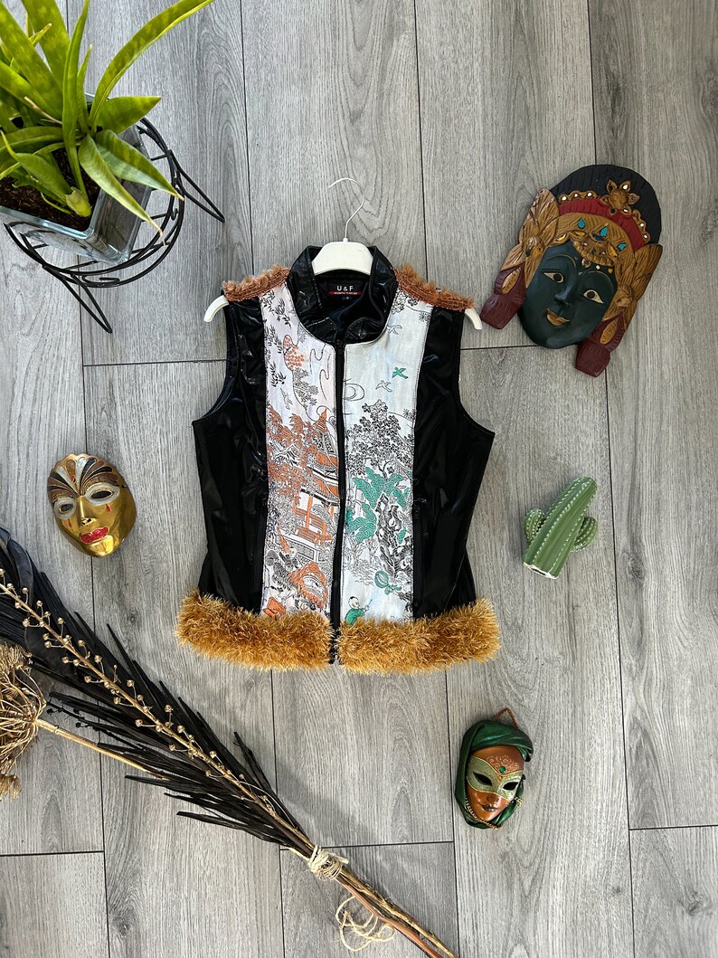 May include: Black sleeveless vest with a white and orange patterned design. The vest has a brown fur trim and a zipper closure. The vest is on a wooden surface with decorative masks and a cactus.
