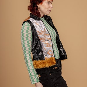 May include: A black shiny vest with a brown fur trim and a white and gold patterned lining. The vest is worn over a green and white patterned long-sleeved shirt and black jeans.