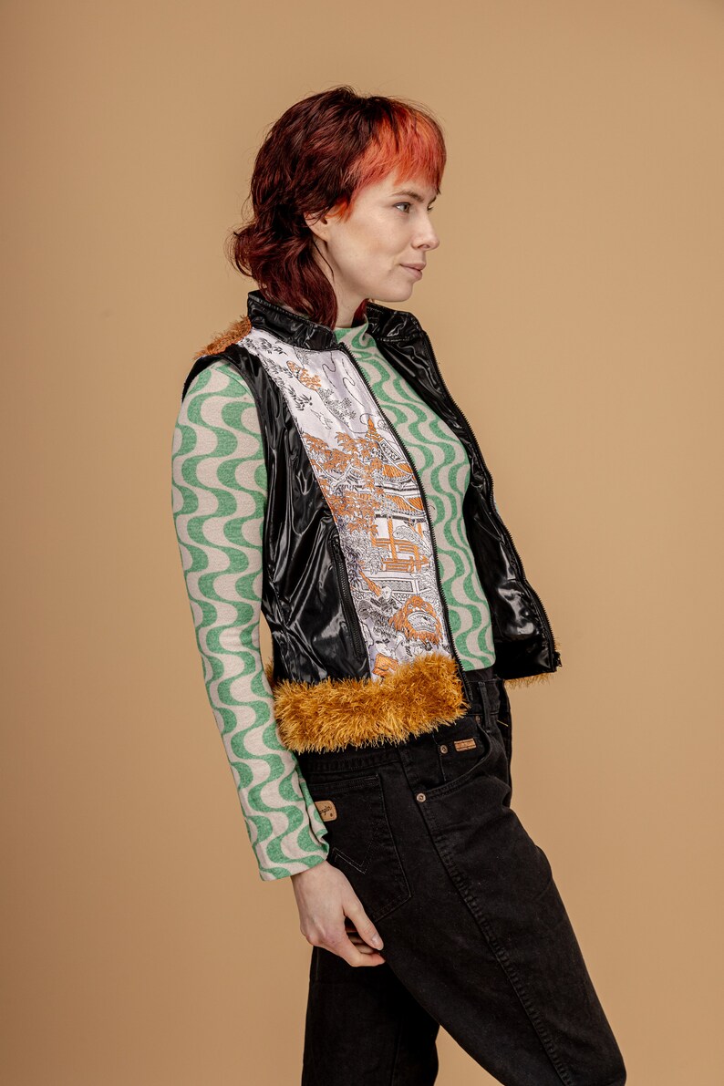 May include: A black shiny vest with a brown fur trim and a white and gold patterned lining. The vest is worn over a green and white patterned long-sleeved shirt and black jeans.